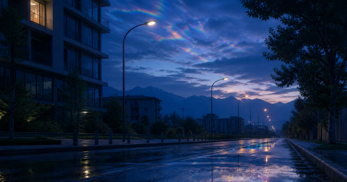 An empty rain-wet street at blue hour, with streetlights and the brightening sky reflected on the pavement