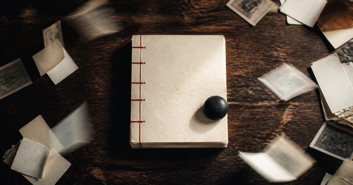 A thick paper volume bound with cinnabar thread lies beneath a black paperweight on a wooden desk while loose pages scatter in the wind