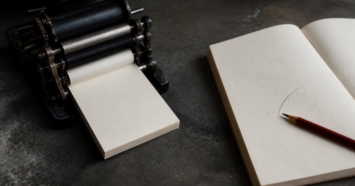A black mechanical paper-feed device produces neat blank sheets while an open notebook beside it holds a single unfinished stroke